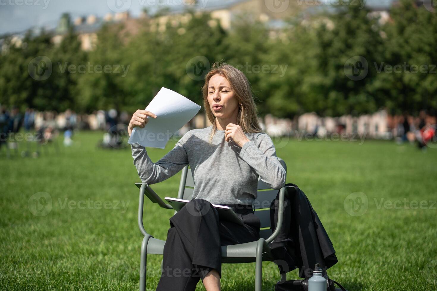 Uncomfortable woman fanning with sheets of paper like fan while sitting on chair outside on lawn. photo
