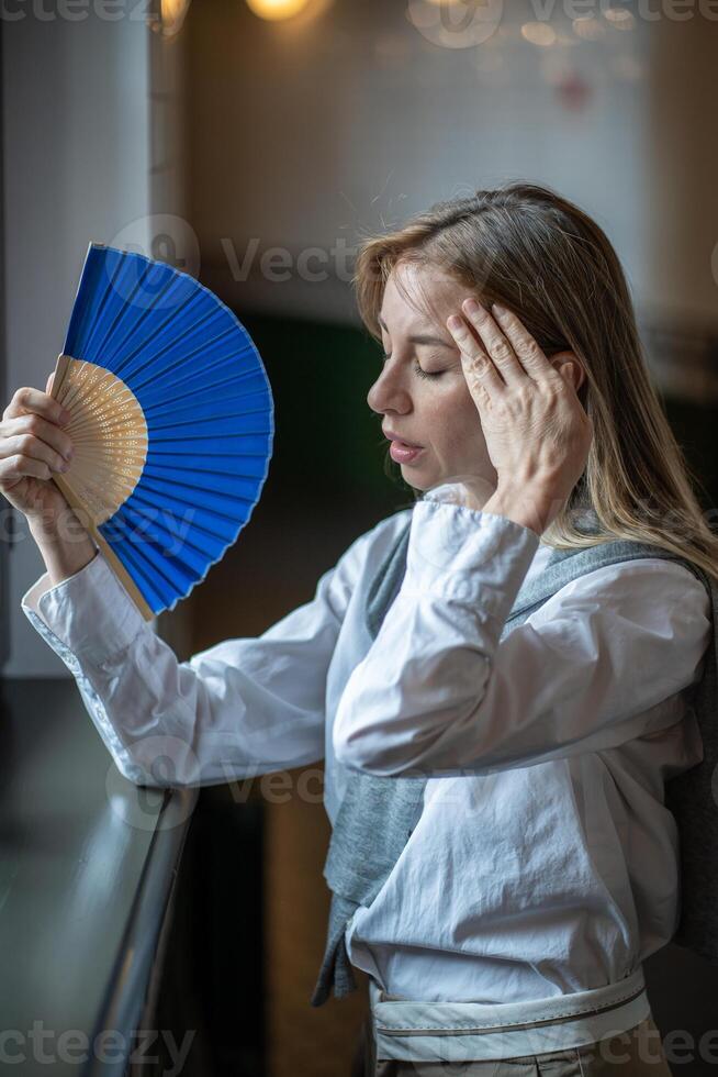 Uncomfortable woman with fan suffers from headache due to high temperature and stuffiness in room. photo