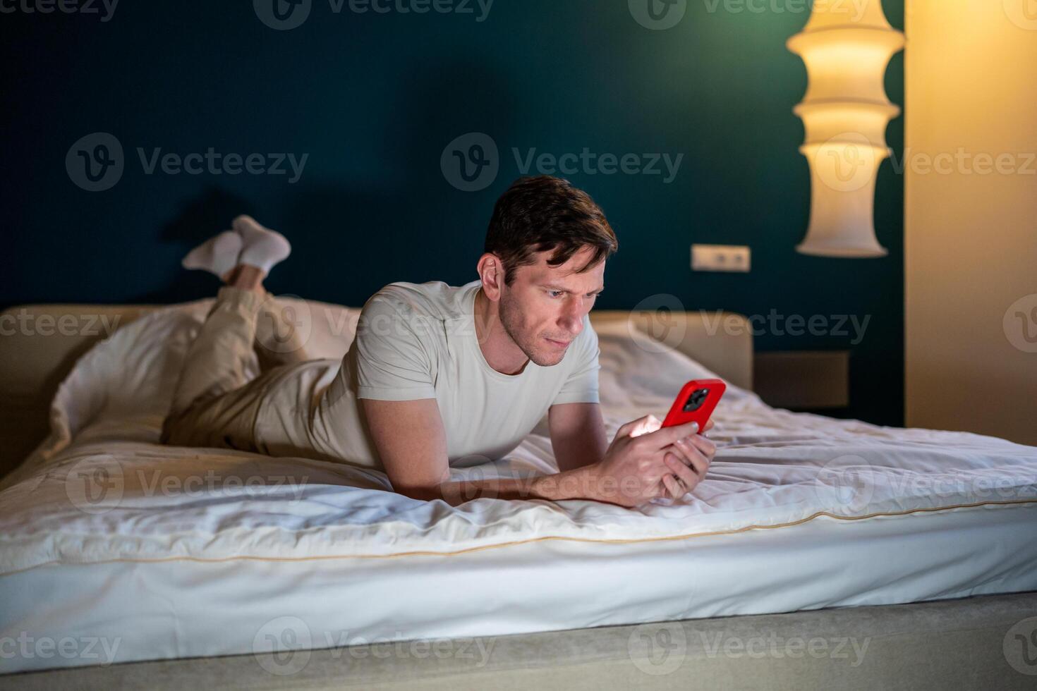 Man reading information in smartphone lying in bed at home in modern bedroom in evening. photo
