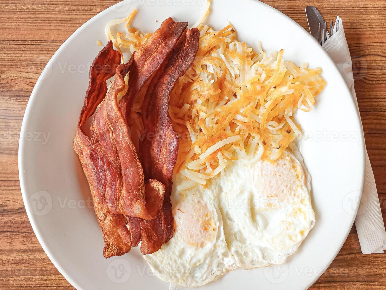 The golden-brown hash browns are piled high on a white plate with crispy bacon strips and a perfectly fried egg nestled alongside. The image evokes the comforting aroma of a classic American breakfast photo