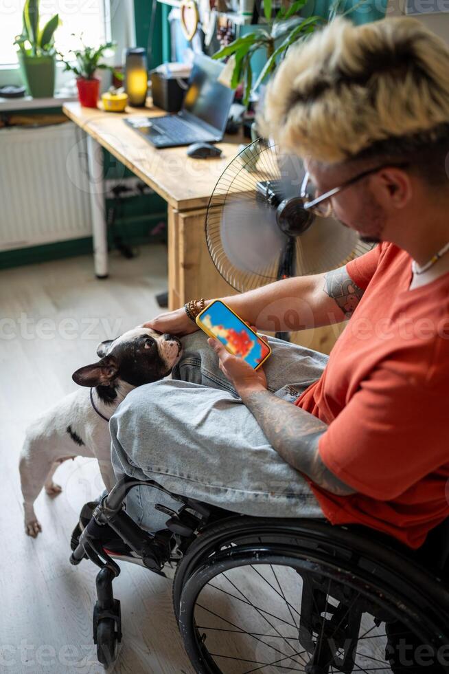 Disabled man in wheelchair looks at weather forecast on phone screen, sits by fan with dog at home. photo