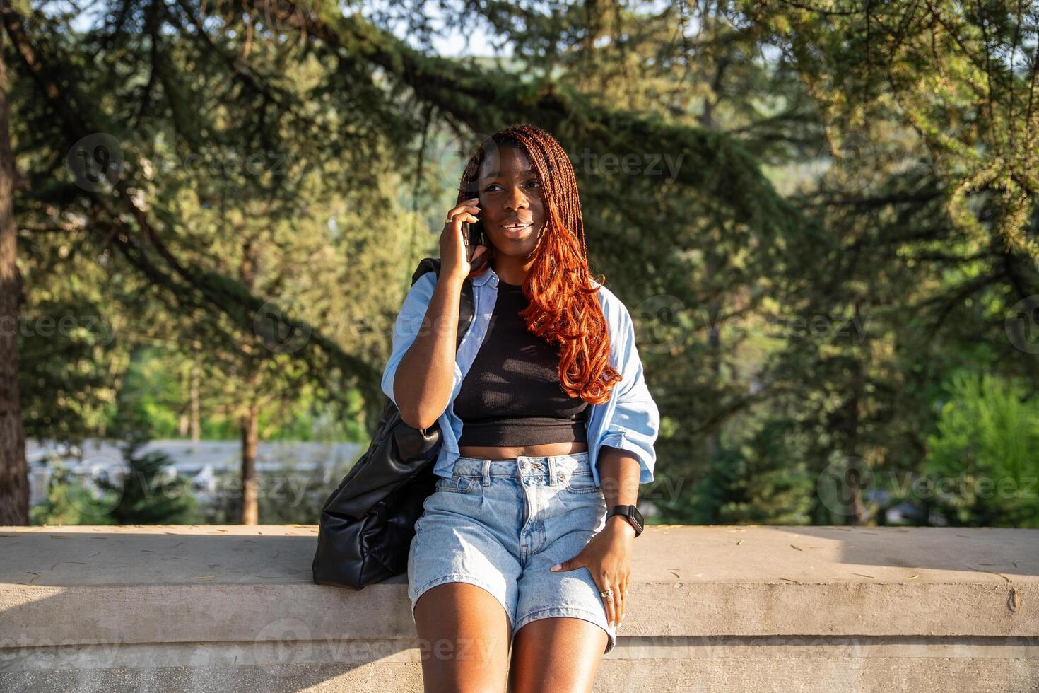 Pleased African American woman taking phone call while walking strolling in park in setting sun. photo
