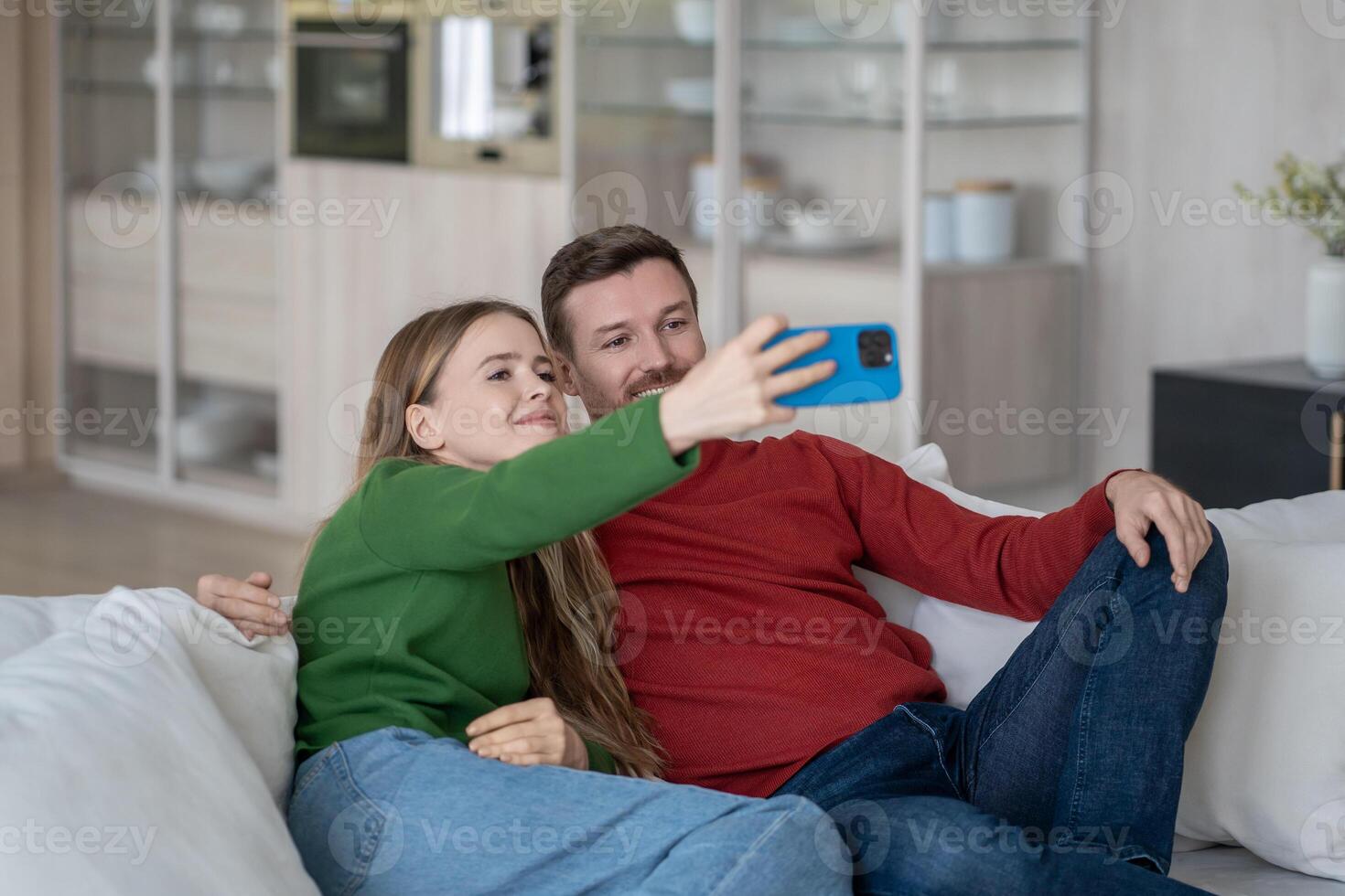 Joyful couple sitting on sofa, looking into smartphone camera, making selfie for sending to parents photo