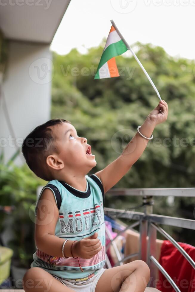 Cute Indian boy holding Indian National Flag in his hangs at home balcony. Bright portrait of happy child sitting on the table with national flag photo
