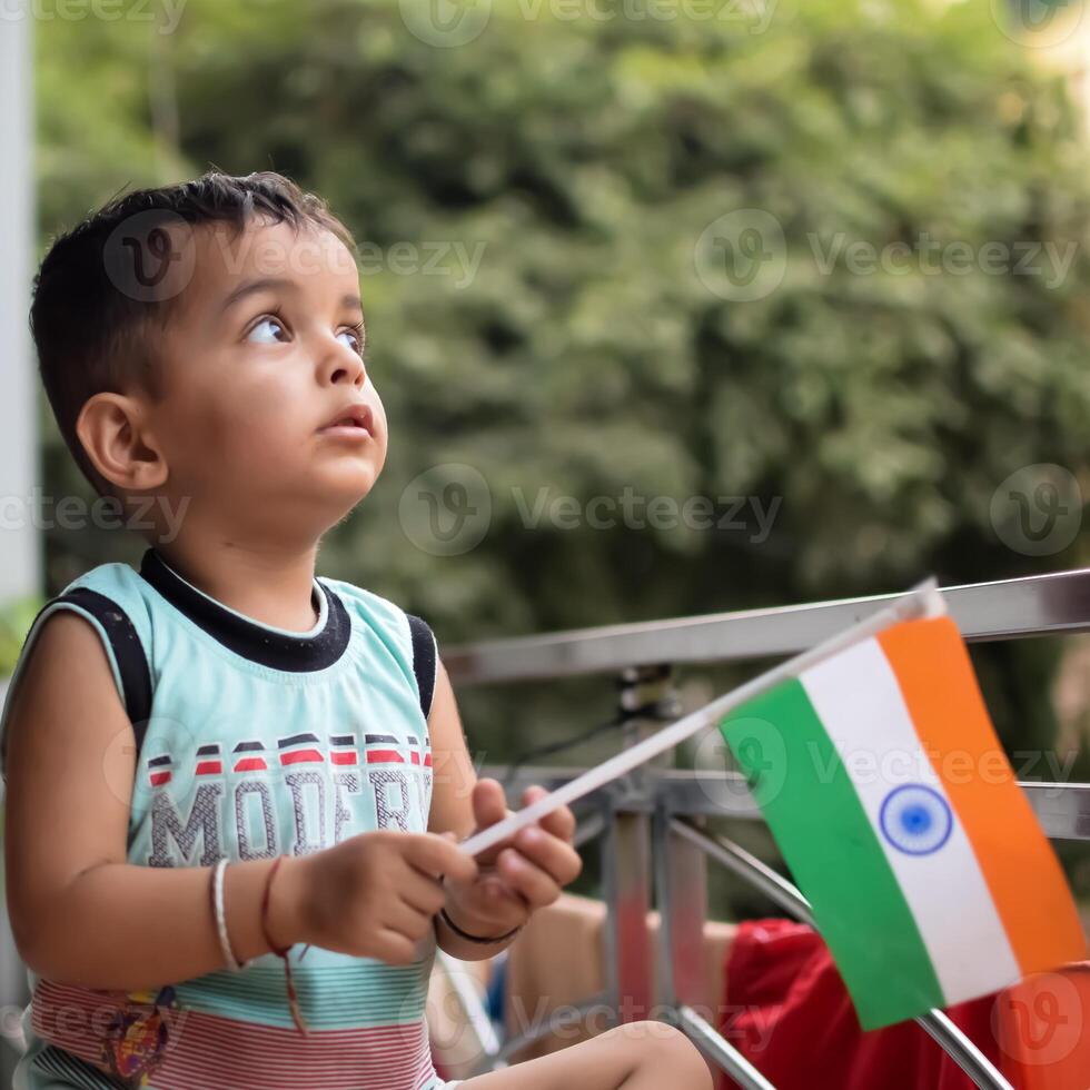 Cute Indian boy holding Indian National Flag in his hangs at home balcony. Bright portrait of happy child sitting on the table with national flag photo