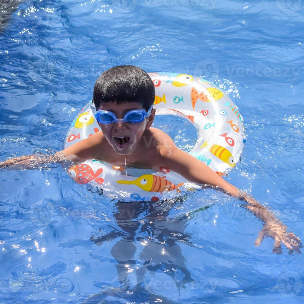 Happy Indian boy swimming in a pool, Kid wearing swimming costume along with air tube during hot summer vacations, Children boy in big swimming pool. photo