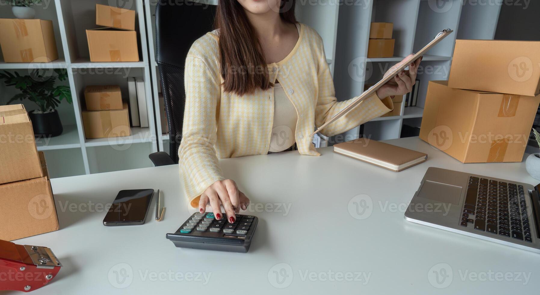 Young Woman Managing Online Sales Orders from Home Office with Calculator and Clipboard, Surrounded by Shipping Boxes photo