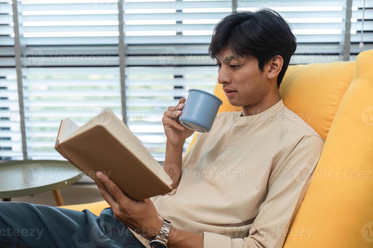 Young Man Relaxing on a Yellow Sofa Reading a Book and Drinking Coffee in a Modern Living Room photo