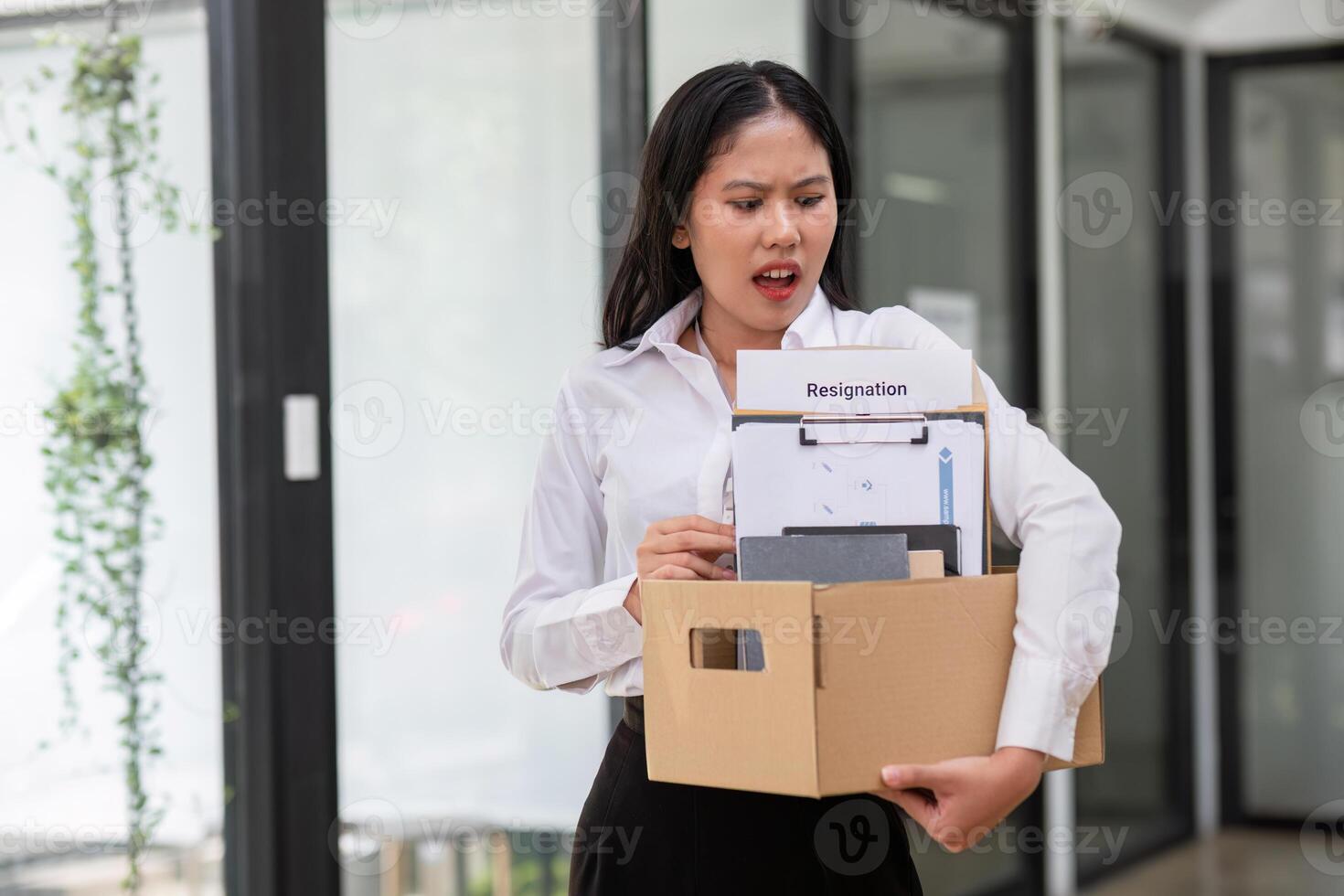Young Female Office Worker Holding a Box with Resignation Document, Expressing Emotion in Modern Office Setting photo