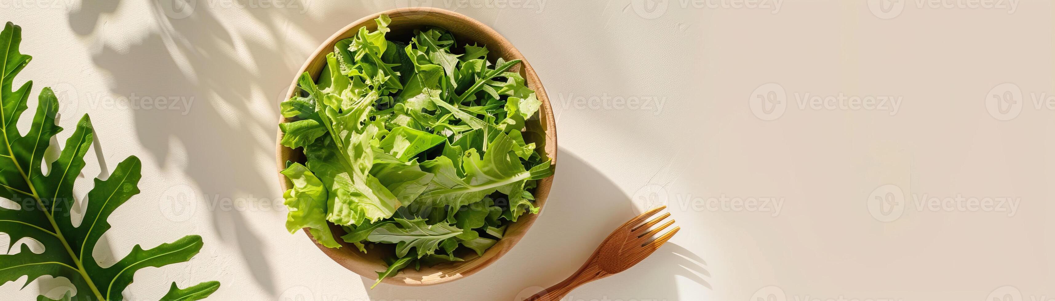 A wooden bowl of fresh green salad on a white table next to a wooden fork. photo