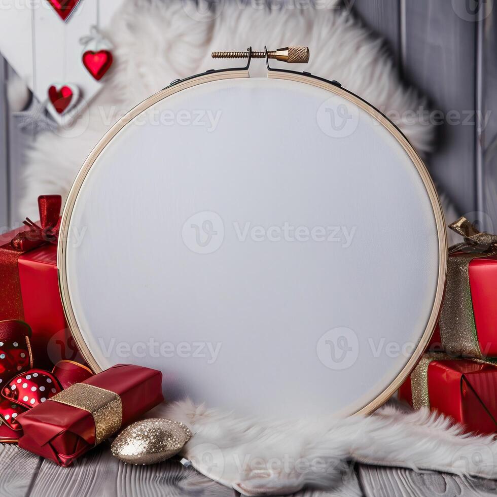 An embroidery hoop with a blank white fabric inside it sits on a gray wooden background surrounded by red and gold Christmas ornaments and gifts. photo