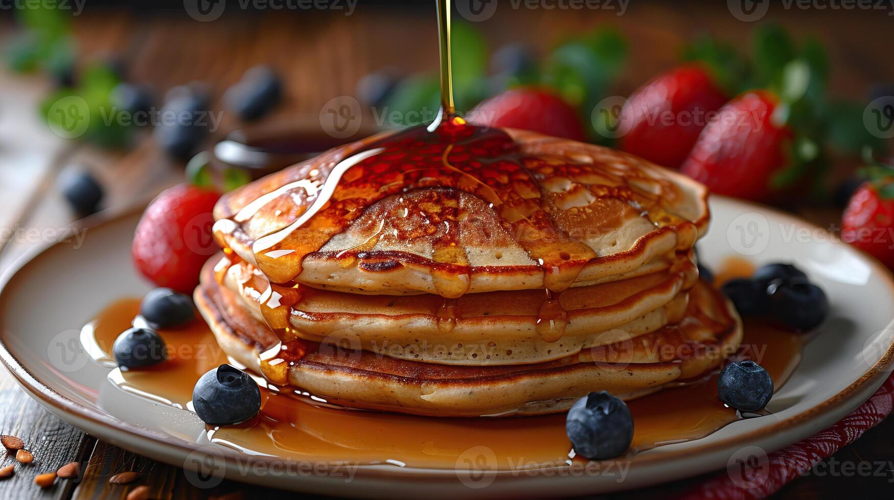 A stack of pancakes with blueberries and strawberries on the side and syrup being poured on top photo