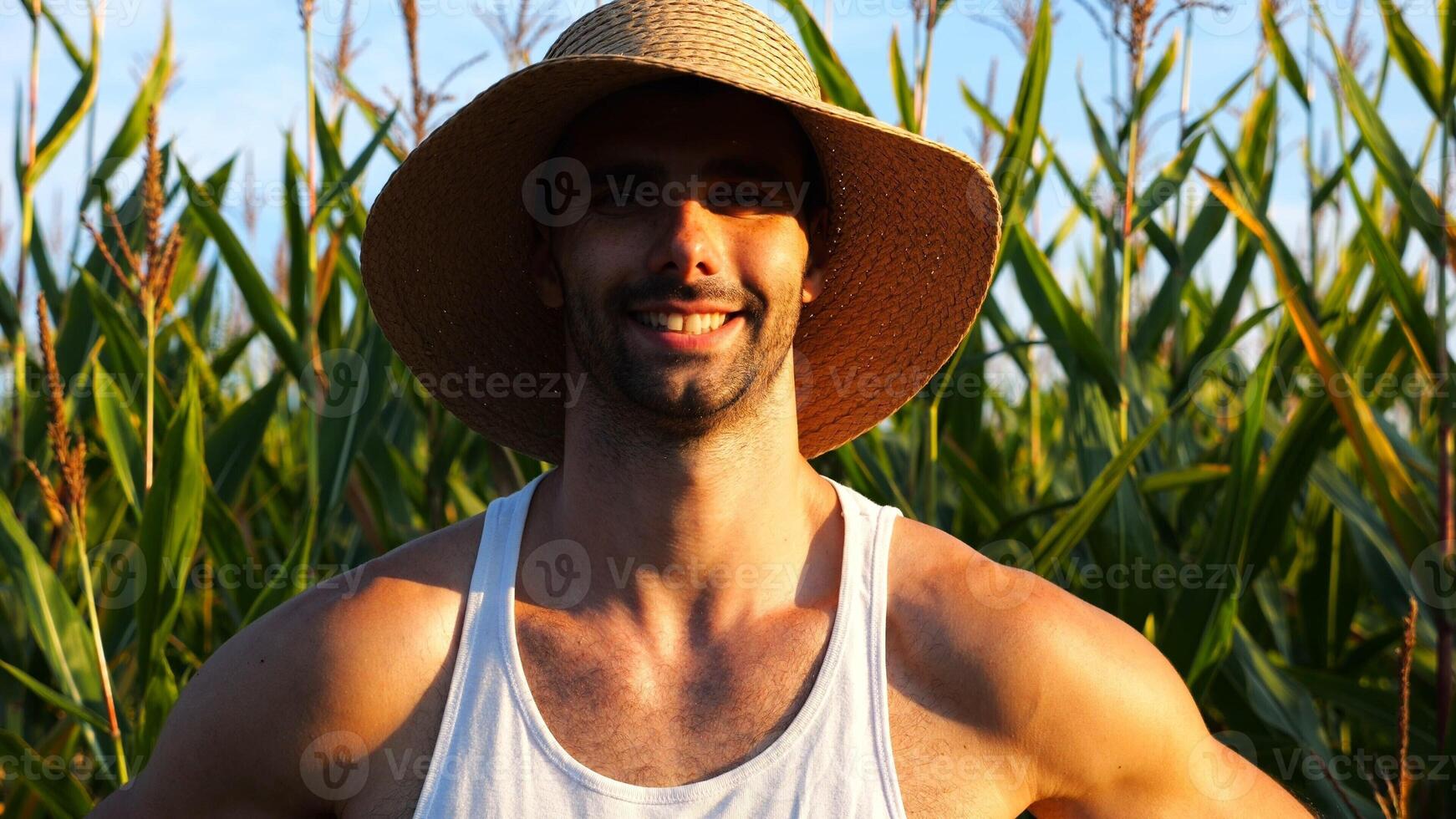 Happy smiling male farmer looks into camera standing near corn meadow. Portrait of young handsome agronomist in straw hat with maize field at background. Agricultural business concept photo