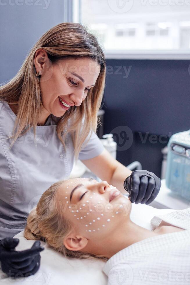 Close up of beautician applies ampoule cream in black gloves to face of female client in spa salon photo