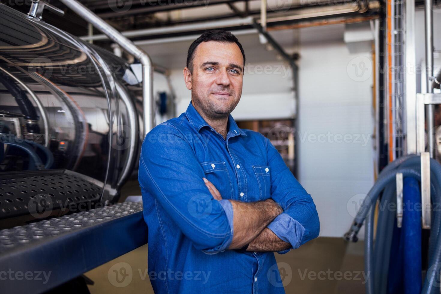A young smiling man in a denim shirt at the cheese factory. An attractive worker near a milk and a butter making machine. An engineer for the maintenance of a cheese production line. photo