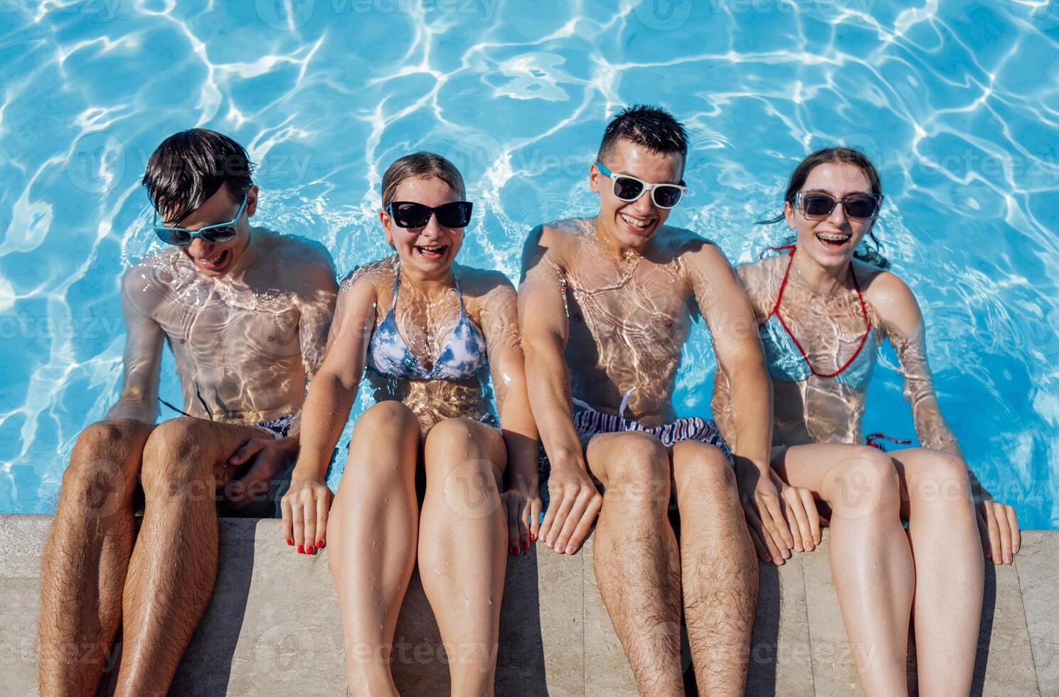Cute teens friends in swimsuits and sunglasses are laughing in the pool. Smiling boys and girls having fun in the clear water. photo