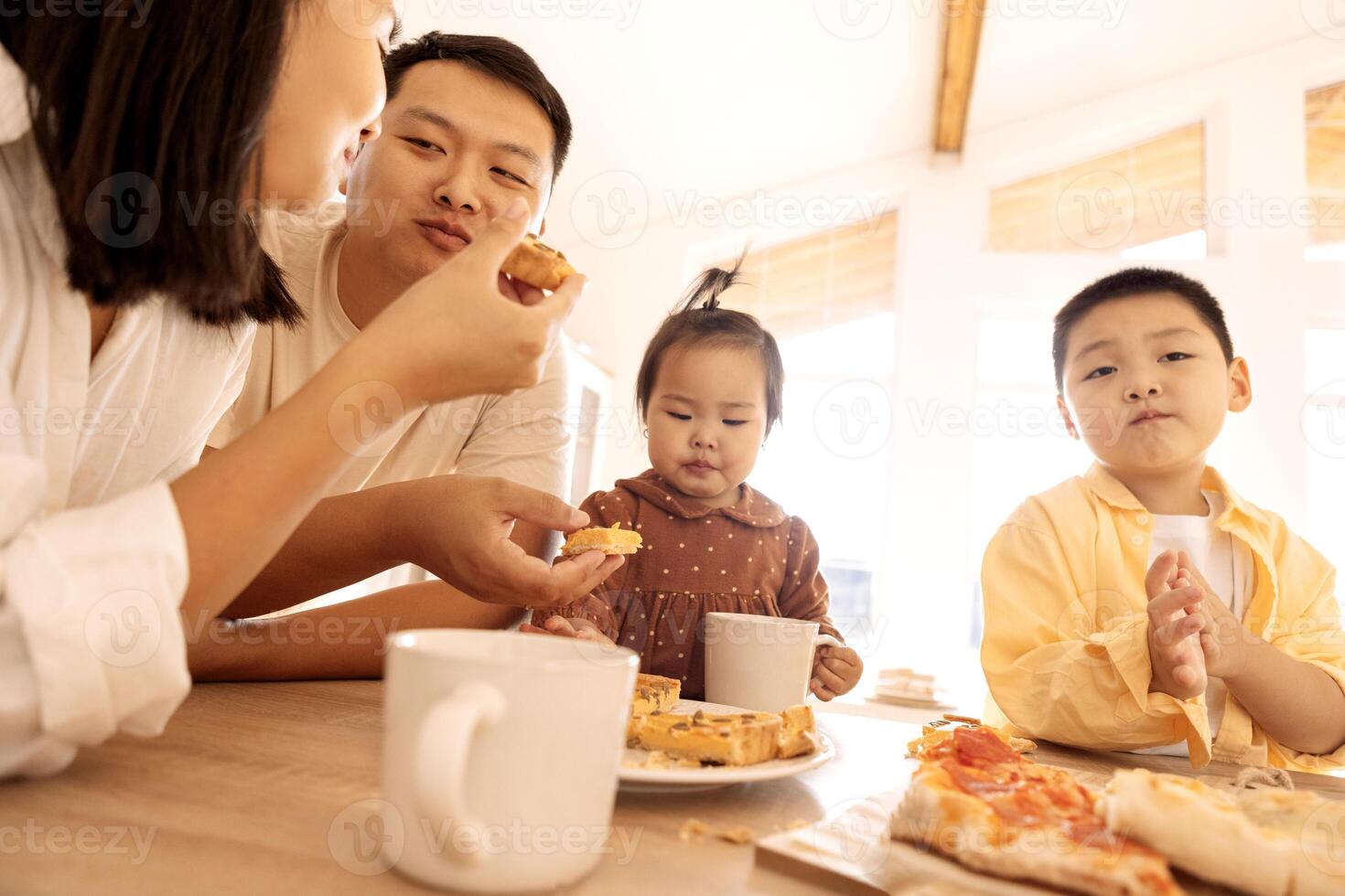 A happy Asian family is eating pizza in the kitchen at home. A young Korean couple and their cute children are having lunch. photo