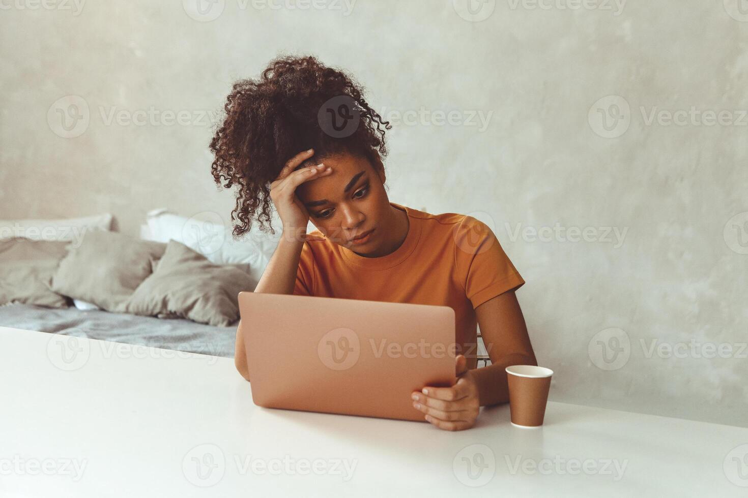 Tired flustrated African descent young girl sitting at desk in front of laptop while irritably looking at computer screen with hands holding her head at temples. photo