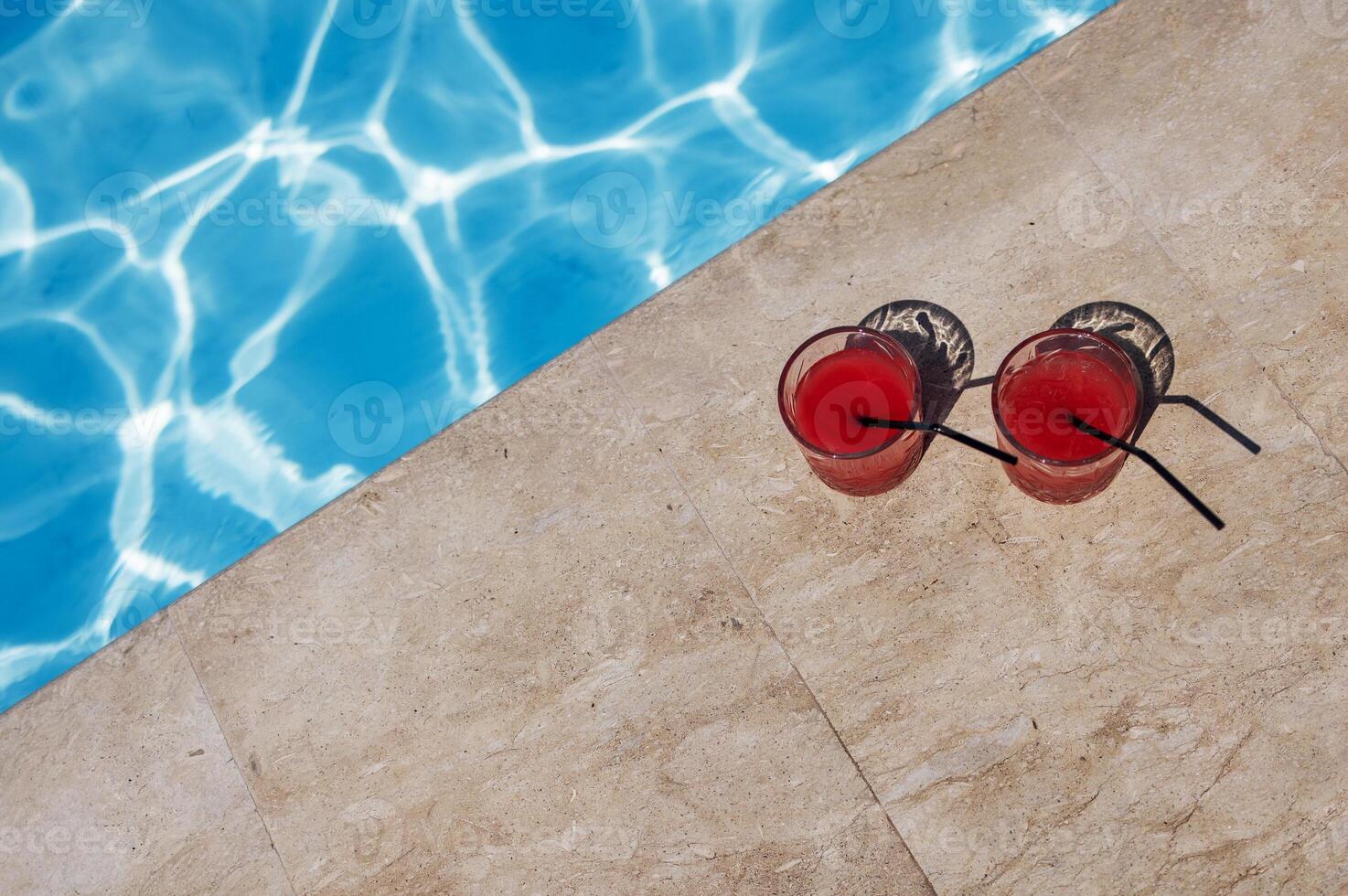 Two glasses with a tasty red cocktail with ice. Delicious fresh drink with straws by the pool. A sunny day and the reflection of rays in the blue water in background. photo