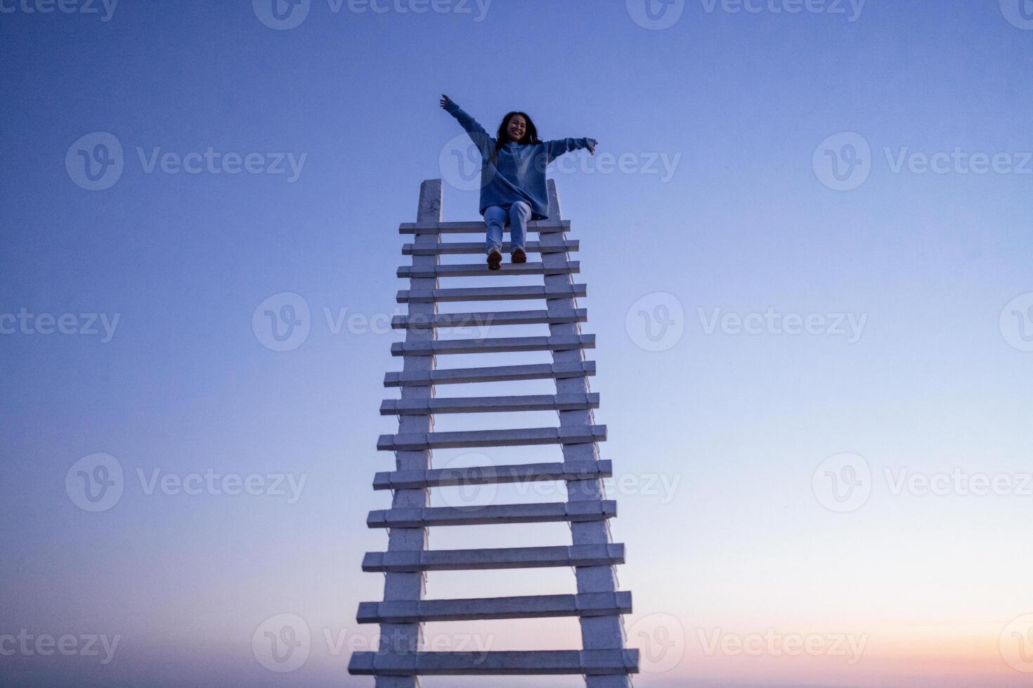 An attractive Asian woman is laughing and sitting at the top of a wooden staircase outdoors. A charming happy Korean woman has spread her arms and is having fun. photo