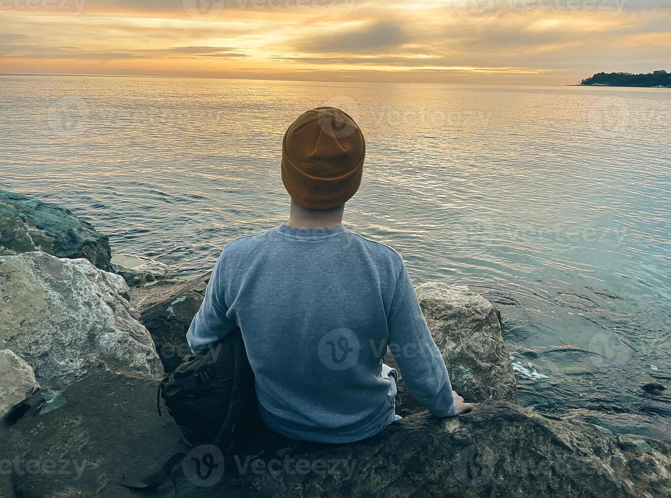 Young man in casual clothes sits on the rocks near the sea. Caucasian guy in a blue sweater and hat holds his jacket and looking at the calm sea. View from the back. photo