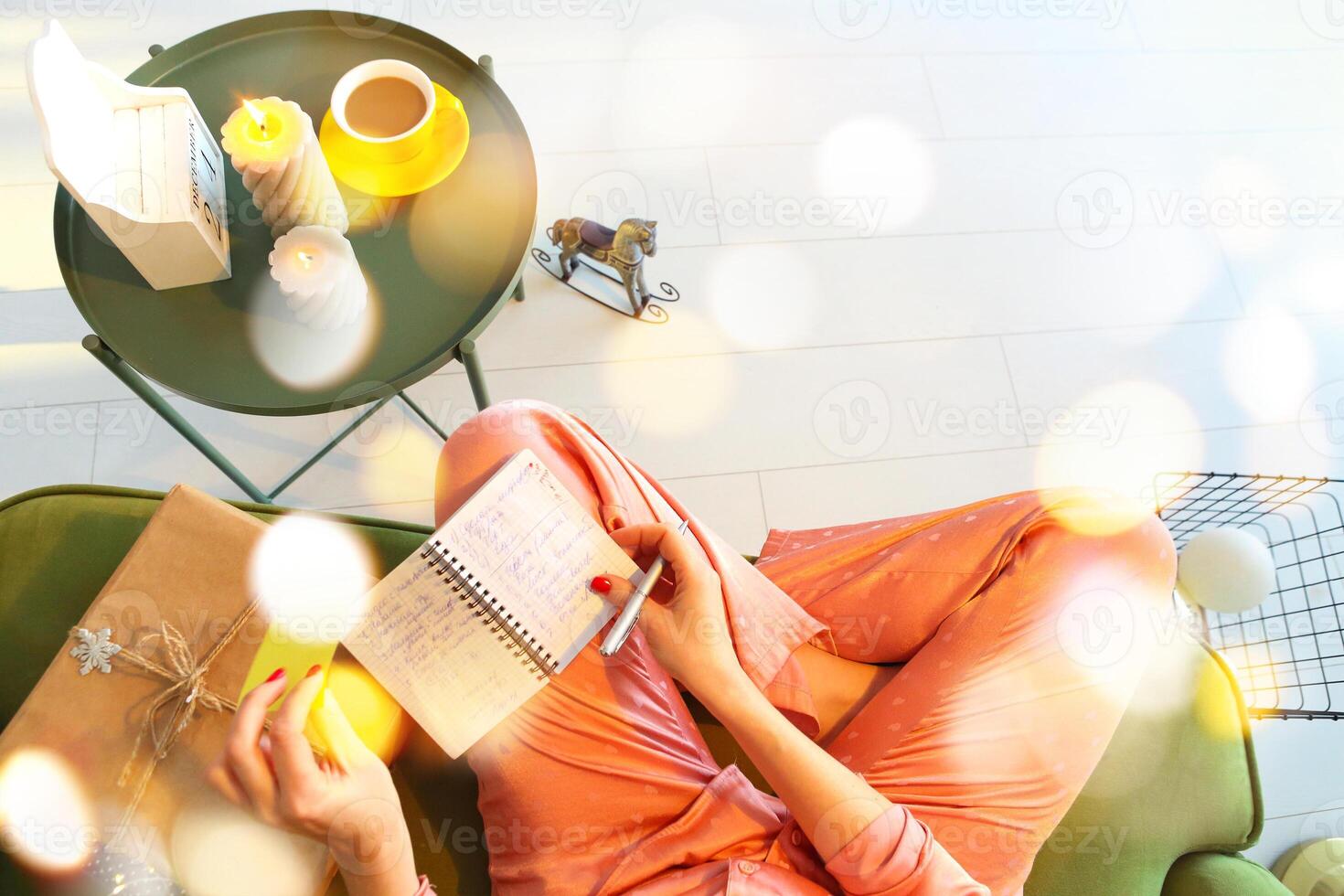 View from above of woman making notes while sitting on sofa with notebook and pen, top view of female wearinng silk pajama planning day or writing todo list during winter holidays. photo