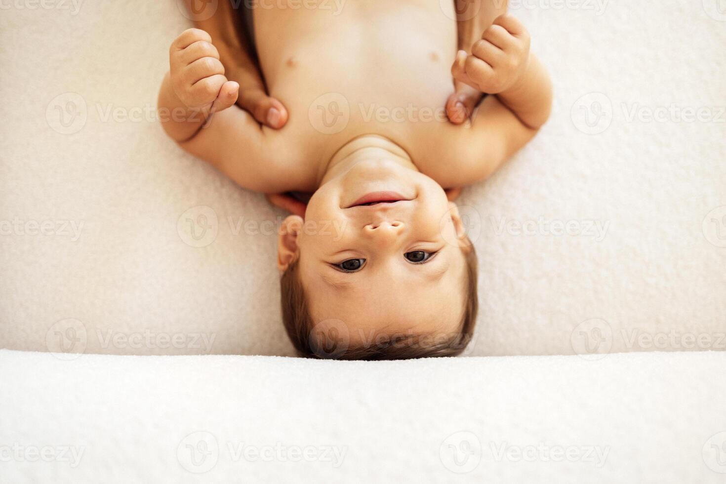 Close-up of the portrait of a cute baby boy. The little sweet toddler is lying on the bed. The smiling child is being held by his mother hands. photo