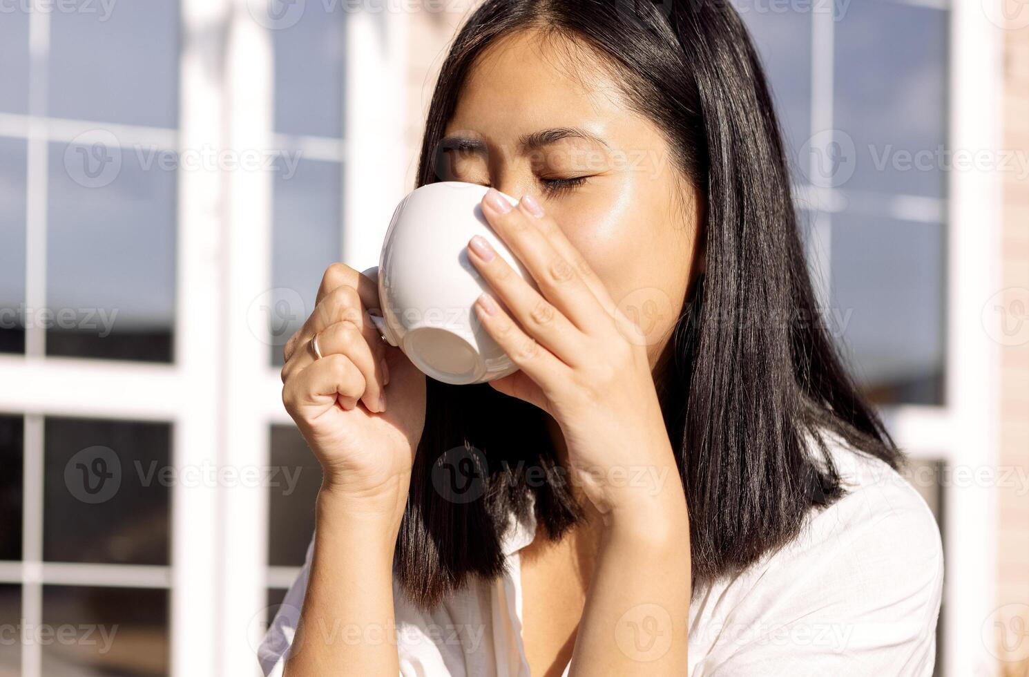 A young cute Asian woman is drinking coffee in the backyard of the house. A charming Korean holds a white cup with her eyes closed. photo