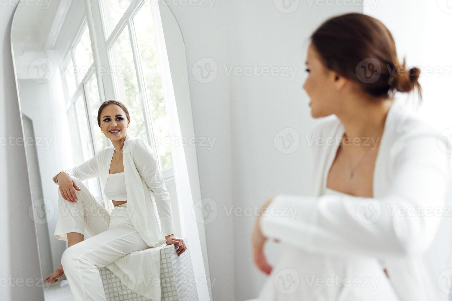 A beautiful girl with evening make-up in white clothes sits on an ottoman and smiles. Reflection of a young woman and panoramic windows in the mirror. photo