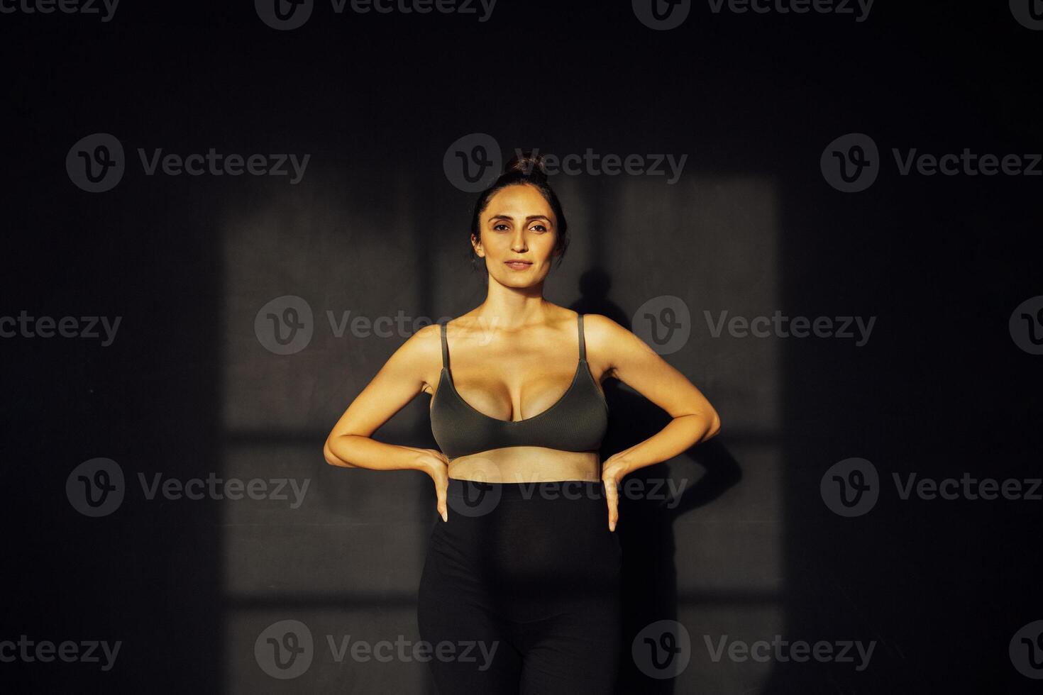 A charming smiling pregnant woman in a dark bra and pants put her hands on her belt and posing in the studio. photo