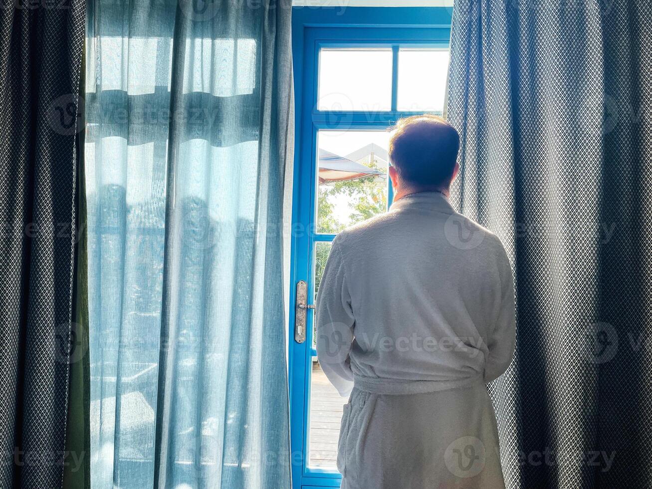 Young man in a white bathrobe looks out the window in his bedroom. View from the back. Green garden is located behand large windows with blue frames and curtains. photo