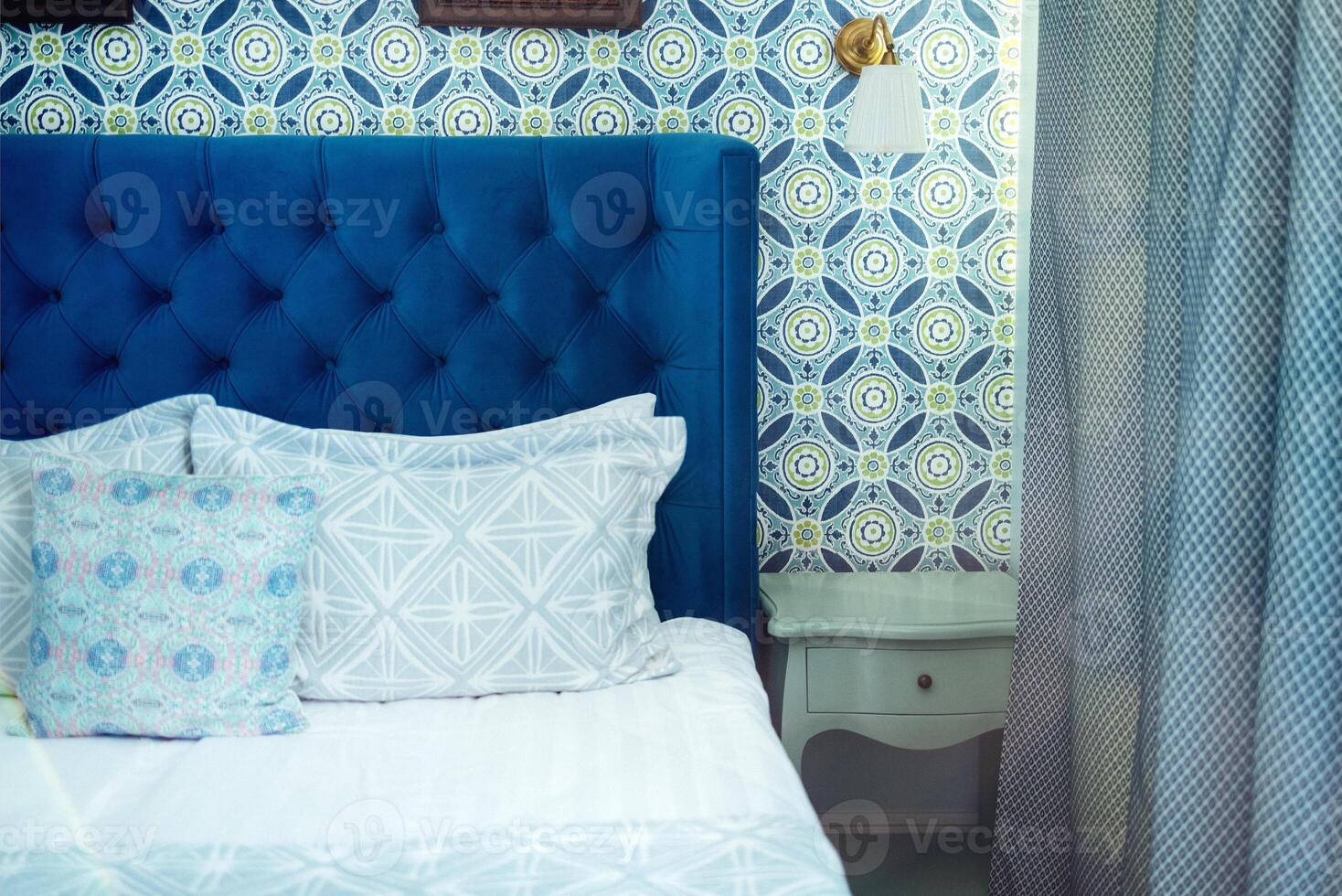 Empty bed with light colored linens, pillows with blue patterns and duvet. Classic wall lamp and wallpaper in bright blue geometric particles. photo