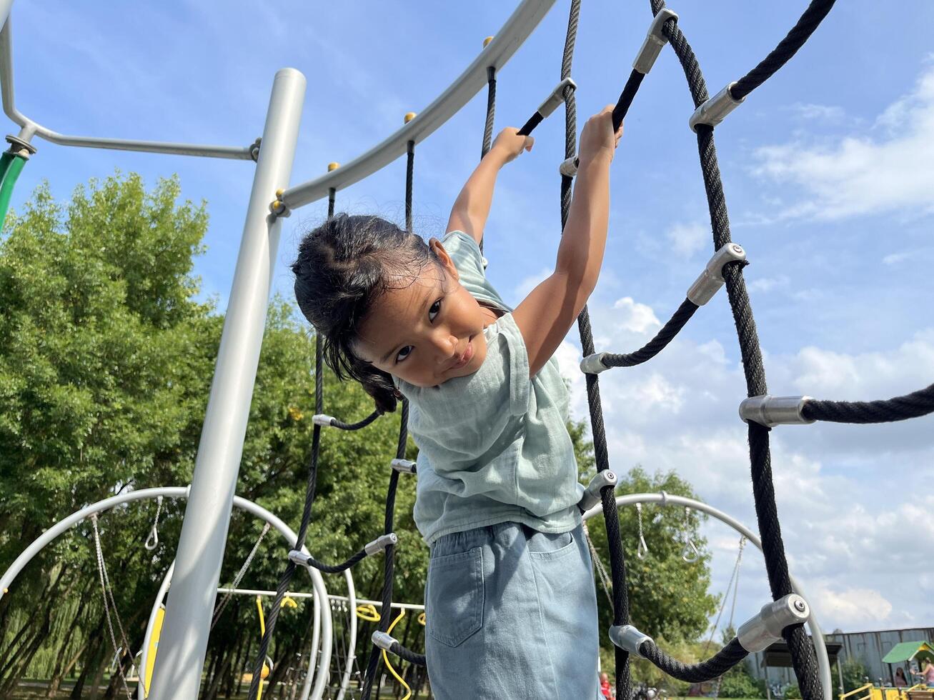 A happy young Asian little girl bravely climbing on a rope in the playground. Childhood concept. Down view photo