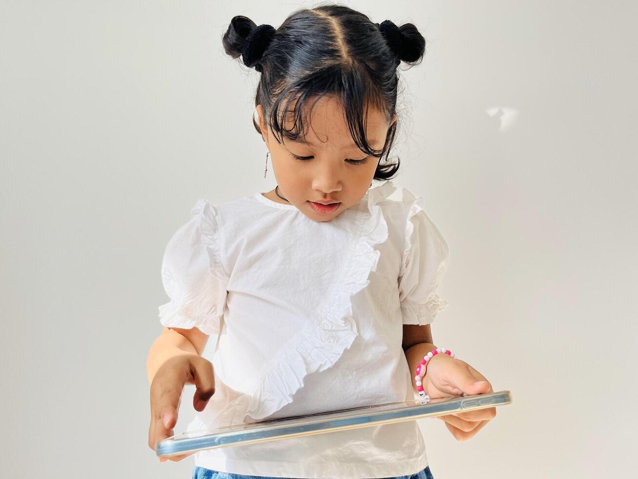 Asian little girl wearing white shirt holding mobile tablet focused watching or playing game with tablet. Gadget addicted concept. photo