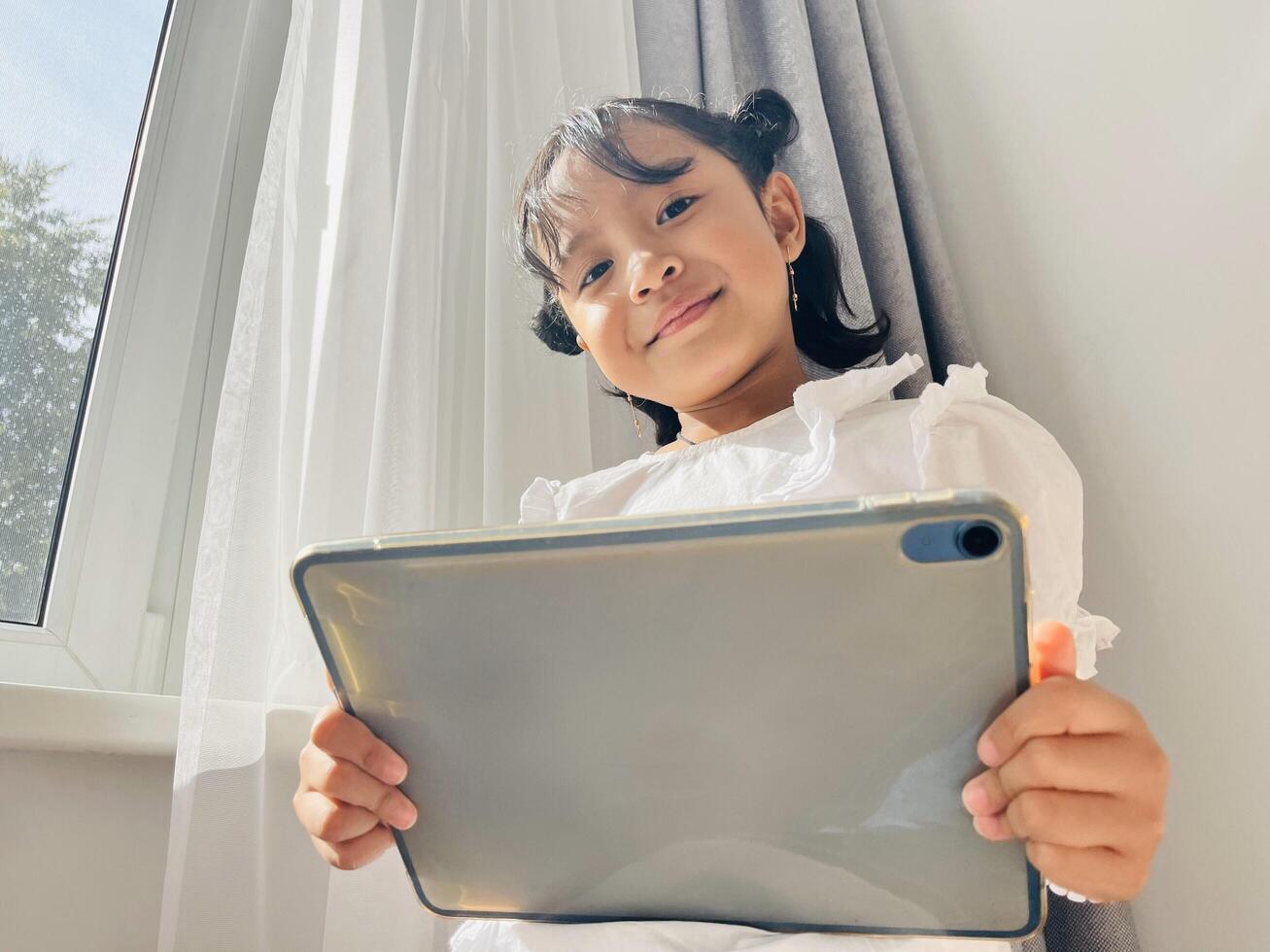Asian little child standing near the window and holding mobile tablet smiling at the camera. Gadget addicted concept. Low angel photo