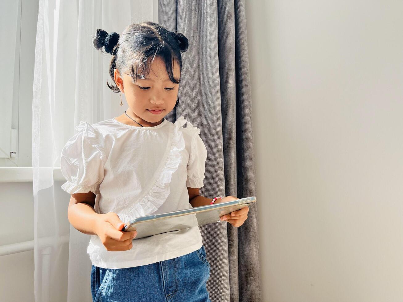 Asian little girl standing near the window holding mobile tablet. Screen time, phone addictive concept photo