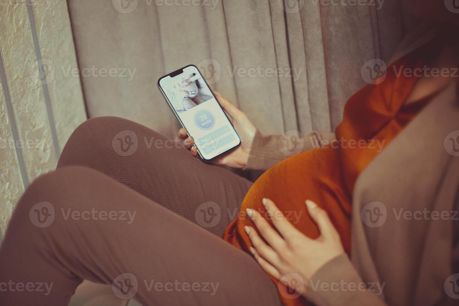 Pregnant woman holding smartphone using pregnancy tracker app on mobile phone while sitting indoors photo