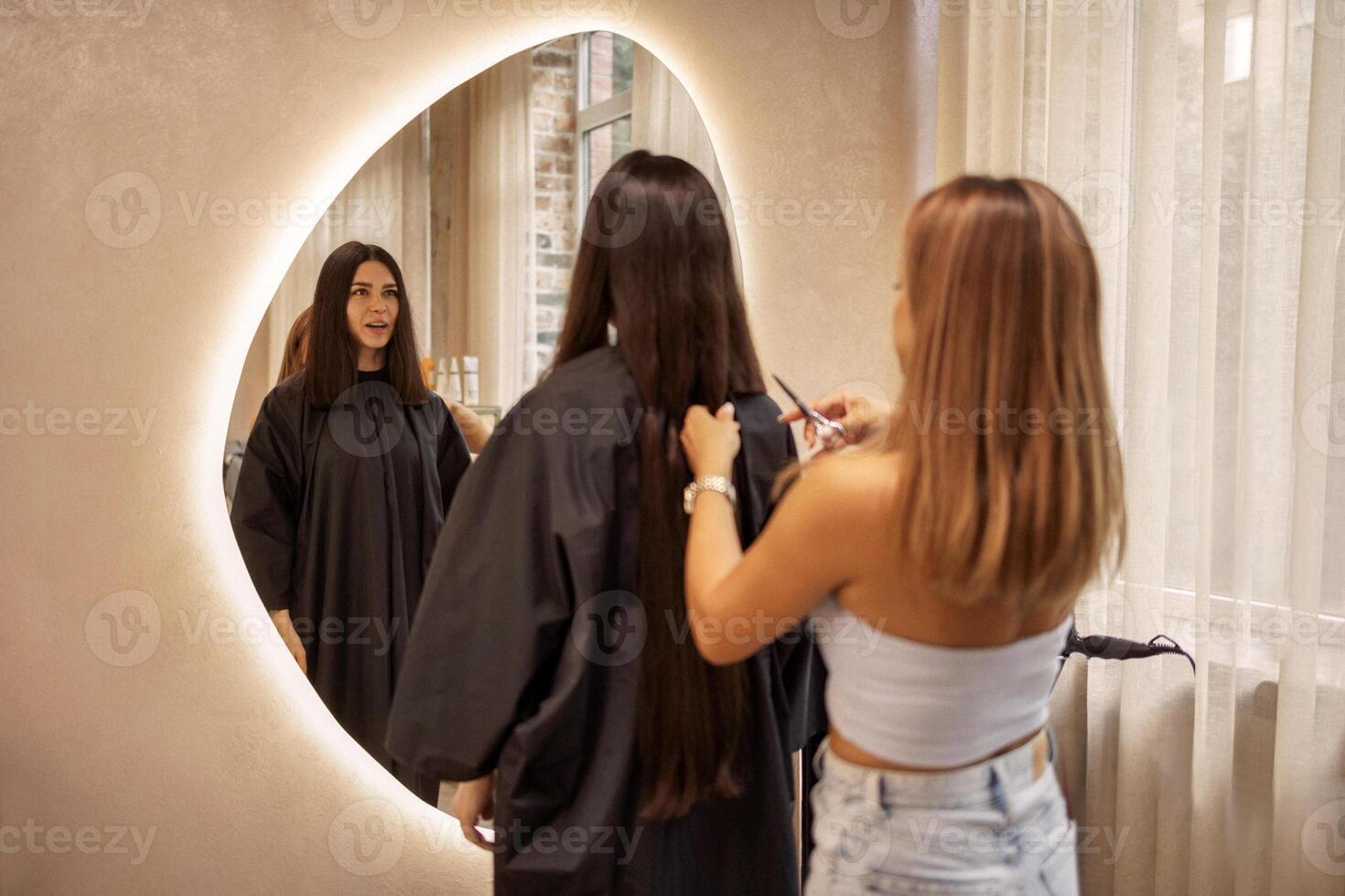 Blonde hairdresser cuts her female client hair in a beauty salon. Pretty brunette young woman looks into a huge mirror when her luxurious long hair is cut off. photo