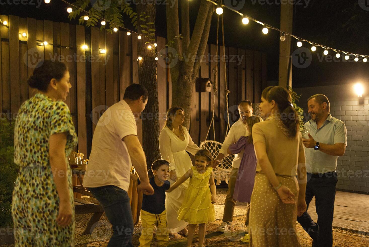 Bride and groom dancing first dance in front of best friends and kids, relatives and guests at illuminated backyard photo