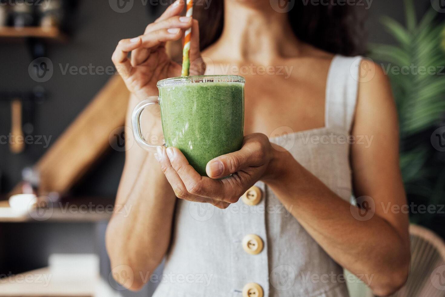 A transparent cup with a green smoothie in the hands of a young woman. A girl in a linen sundress drinks a fresh drink made from herbs and vegetables in the kitchen. photo