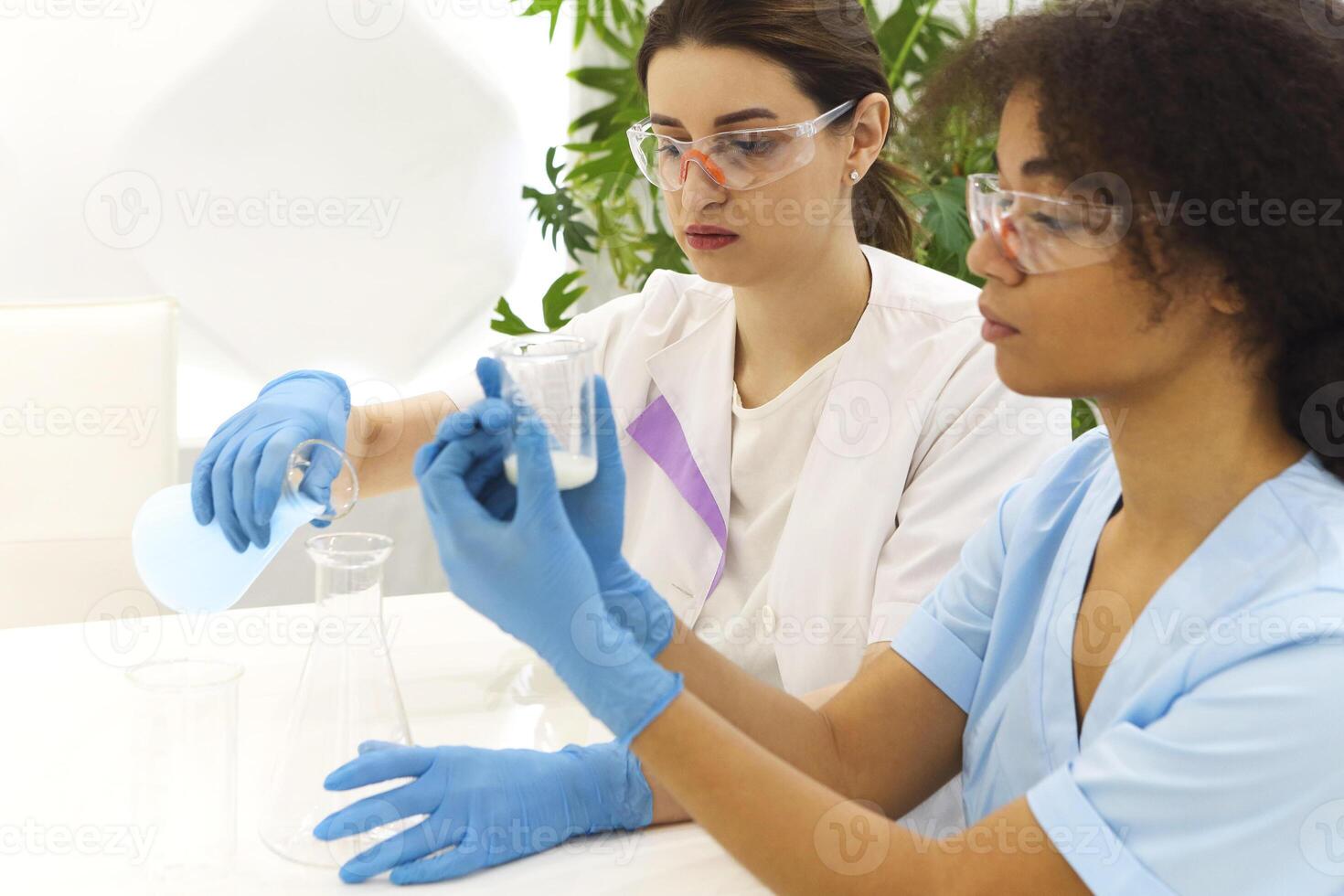 Two multi-ethnic female lab scientists in protective uniform carrying out experiments in laboratory photo