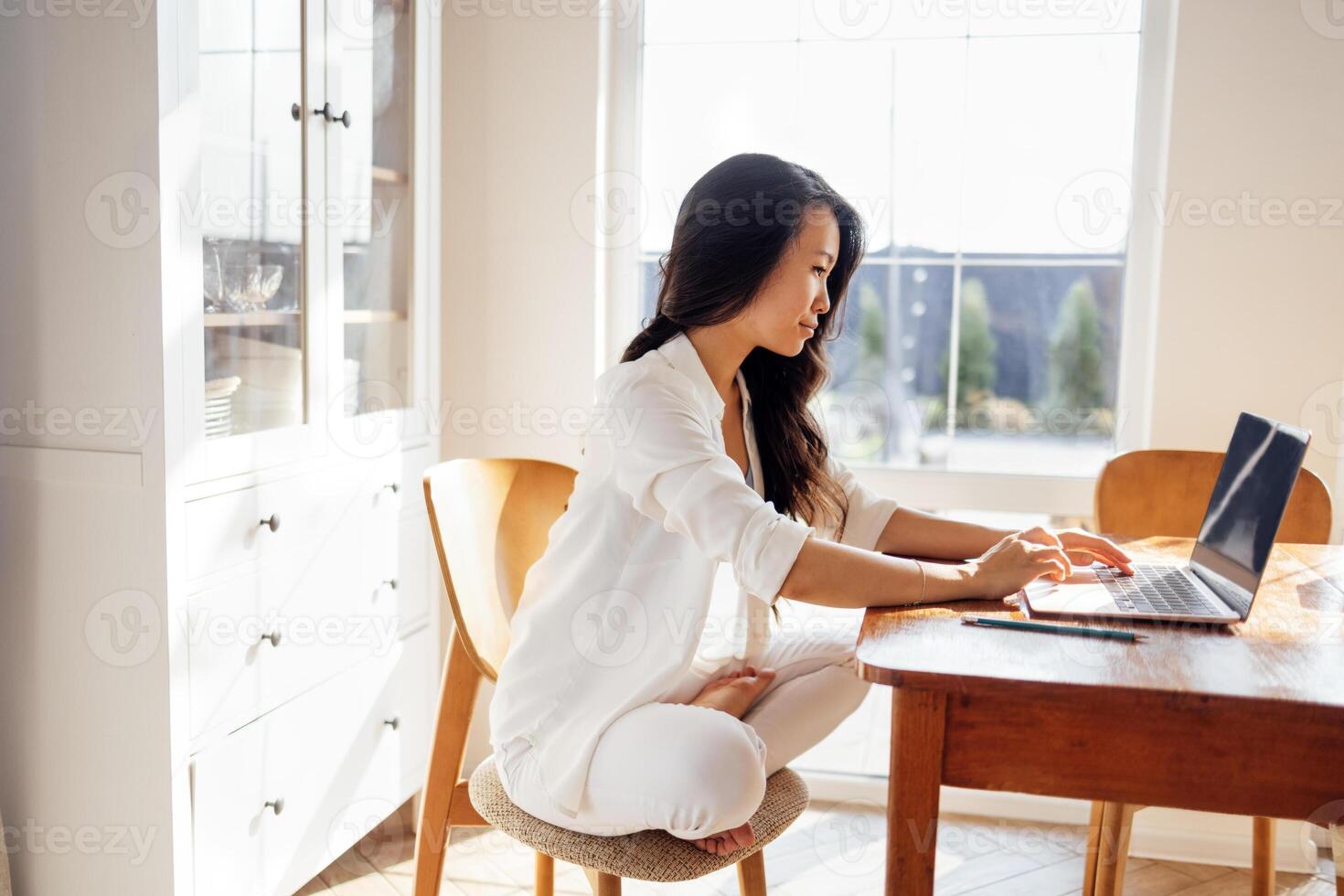 Young charming Asian woman is working at table with laptop in dining room. Lovely Korean girl is studying online at home. Attractive businesswoman communicates via link. photo