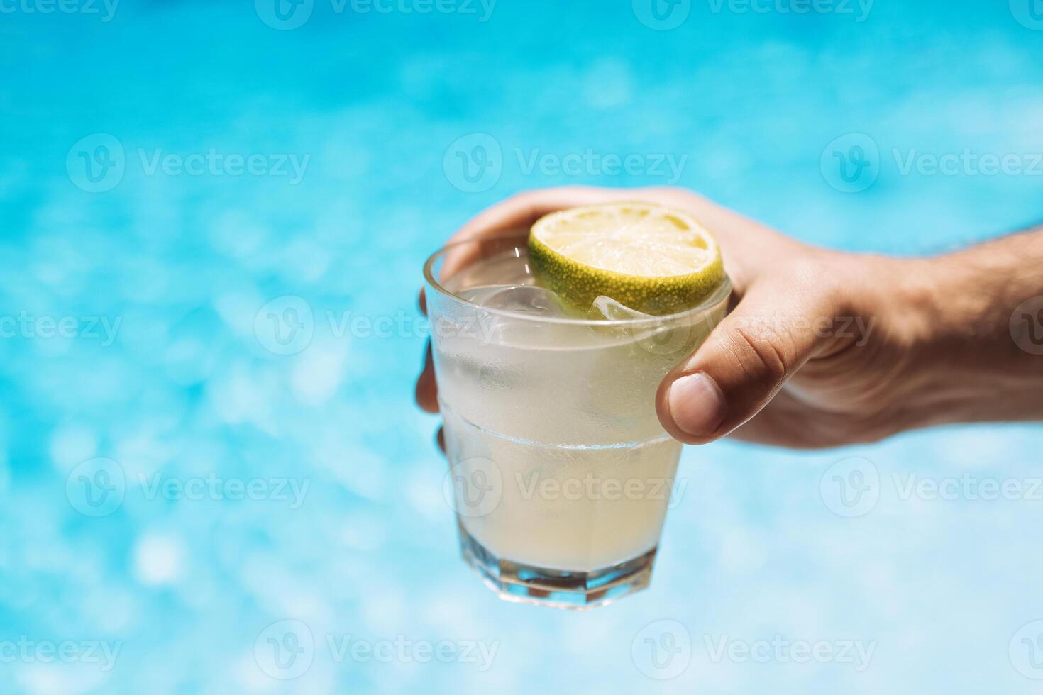 Young male hand holds a glass of a chilled drink with ice cubes. White cocktail with a lemon slice. Sunny day and reflection of rays in the blue pool water on background. photo