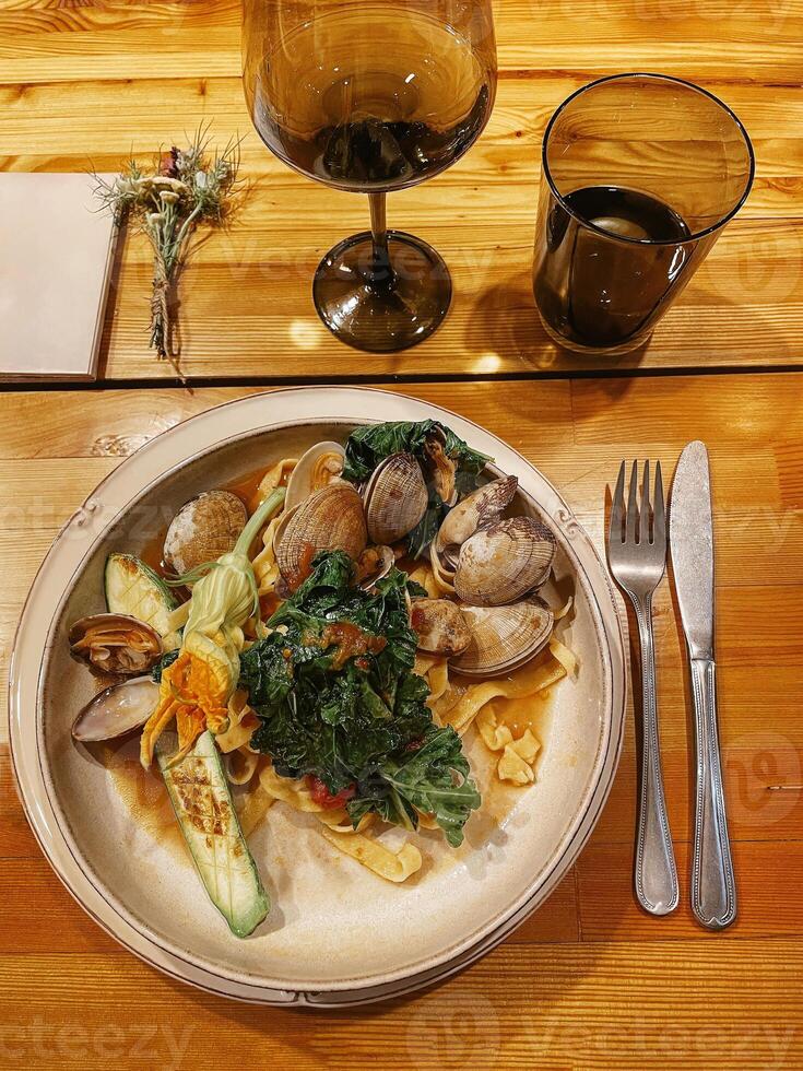 Gourmet lunch with mussels, pasta, zucchini and greens. The dish is served with a fork, knife and glasses. Next to it lies a menu and a bouquet of flowers. photo