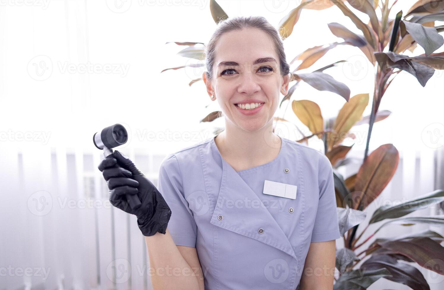 A young woman in a doctor uniform and black gloves with a dermatoscope in her hand in a beauty salon. photo