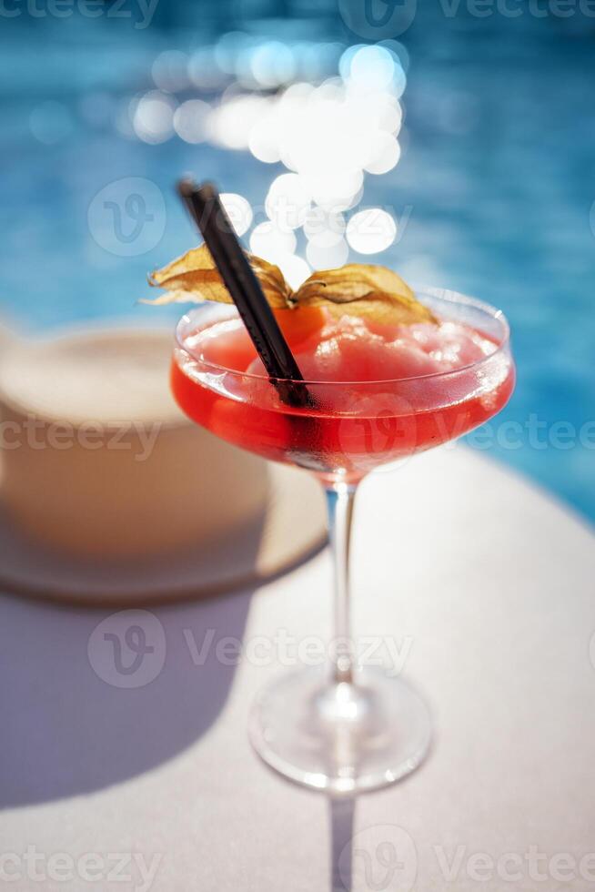 A red drink in an elegant glass with a long stem on the side of the pool. An alcoholic ice drink decorated with a flower on a background of turquoise water. photo