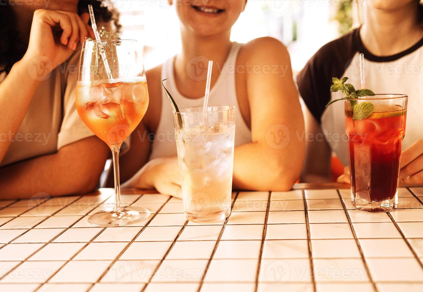 Close-up of three charming girls drinking chilled cocktails in a cafe. Glasses with delicious fruit drinks and ice cubes. Smiling friends relax and drink lemonade in a restaurant. photo