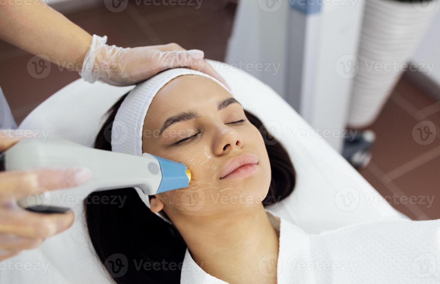 Cosmetologist in grey uniform and transparent gloves makes facial cleansing procedure for her young client photo