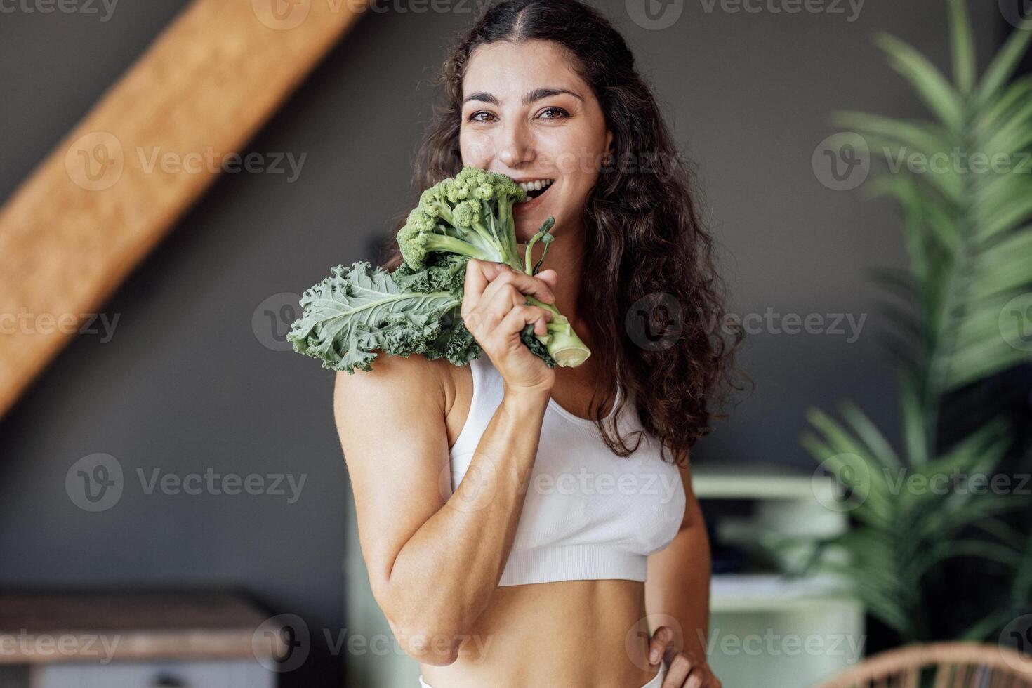 An attractive smiling vegan girl in a white top bites fresh broccoli. A curly-haired young fit woman laughs and eats cabbage at home. photo
