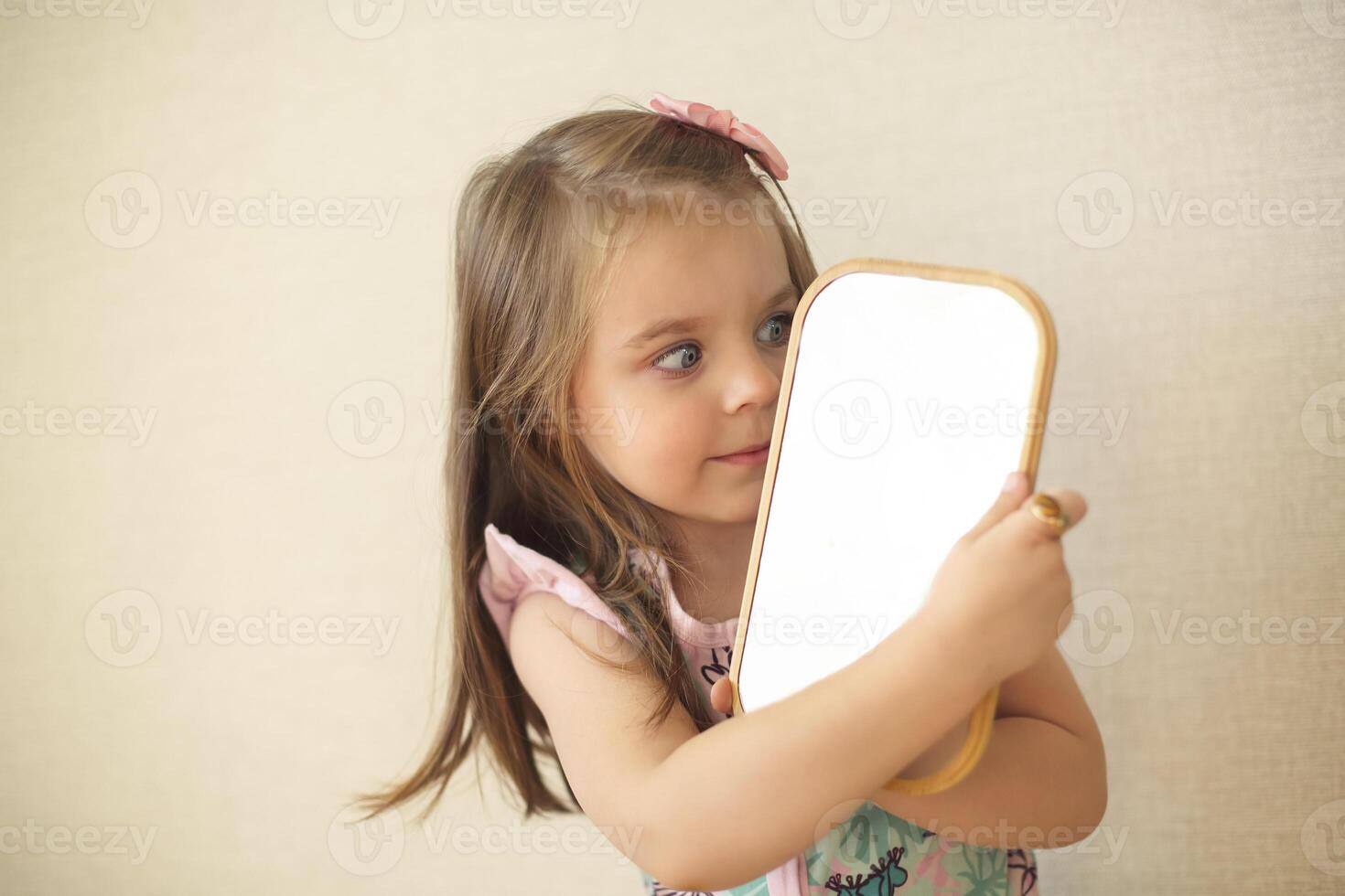 Young curious little girl holding mirror and looking at her reflection while playing alone at home photo
